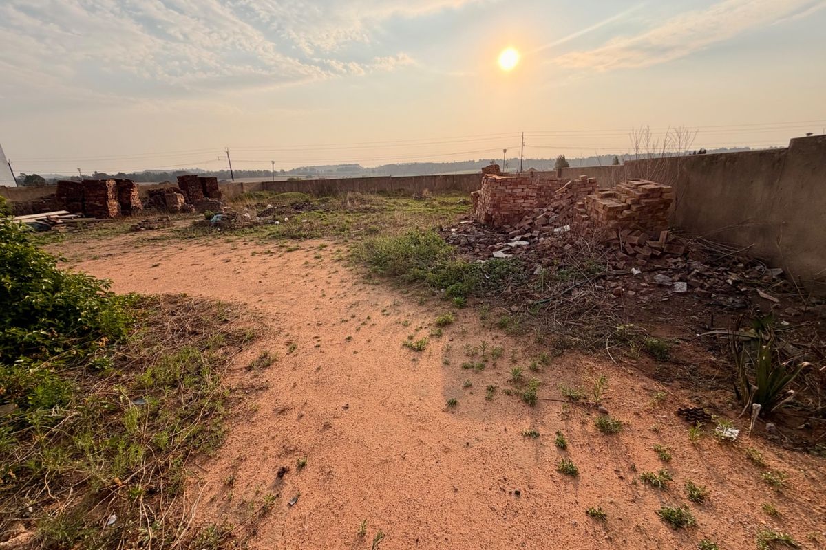 A vacant residential plot featuring a boundary wall, sandy terrain, and scattered construction debris under a sunset sky.