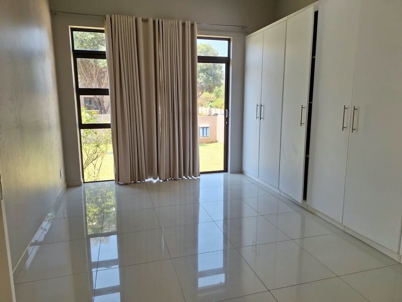 A bright, empty bedroom featuring modern white built-in cupboards, polished tiled flooring, and large aluminium-framed windows with access to the garden.