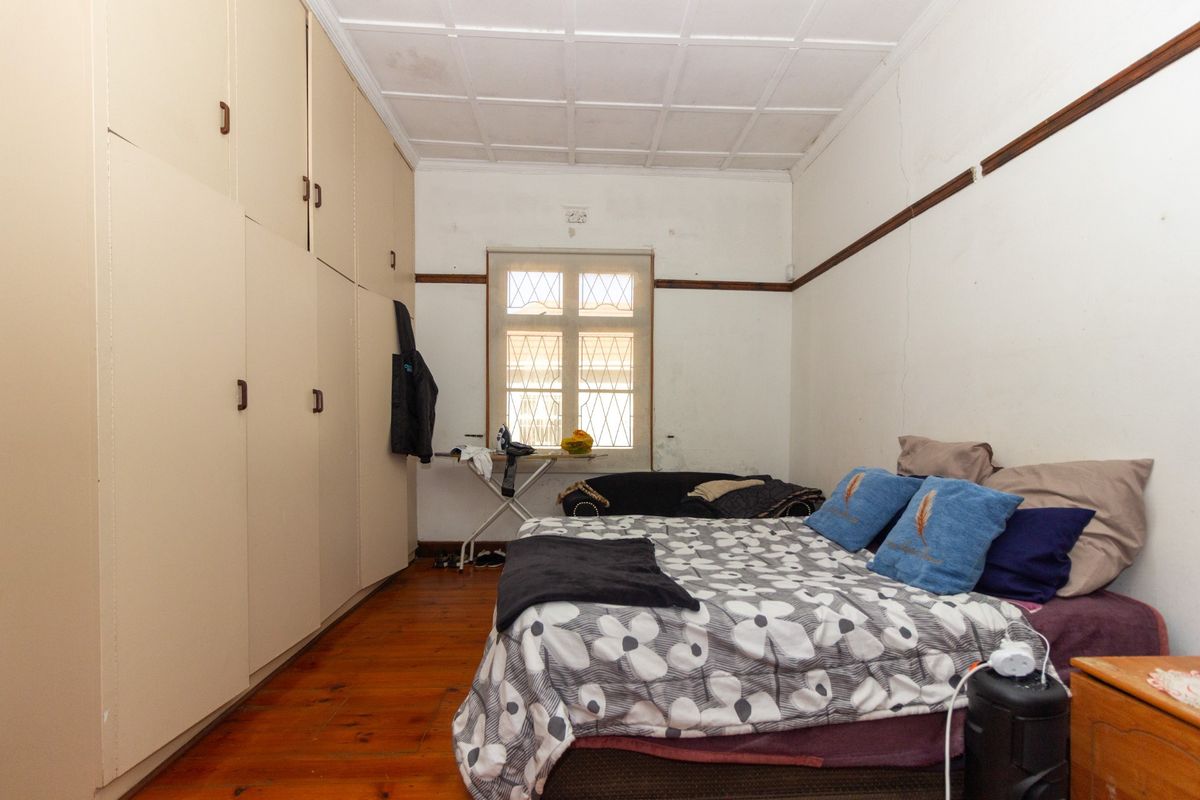 Bedroom featuring wooden flooring, built-in cupboards, and a window with security bars. The room requires cosmetic updates to the walls and ceiling.