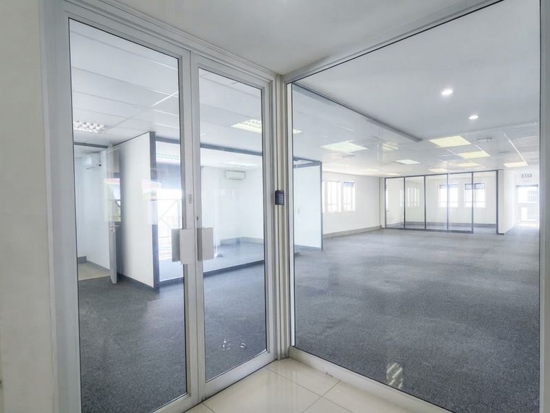 A bright, open-plan office space featuring glass partitions, suspended ceilings with recessed lighting, and carpeted flooring, offering a clean and professional work environment.
