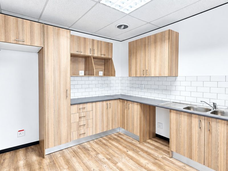 A clean, modern office kitchenette featuring light wood-grain cabinetry, subway tile backsplash, and a stainless steel sink with ample storage space.