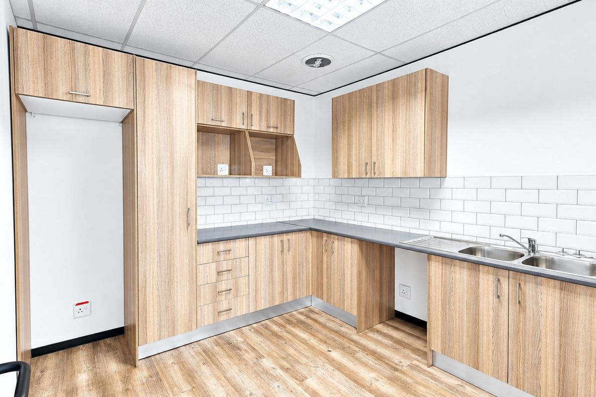 A clean, modern office kitchenette featuring light wood-grain cabinetry, subway tile backsplash, and a stainless steel sink with ample storage space.
