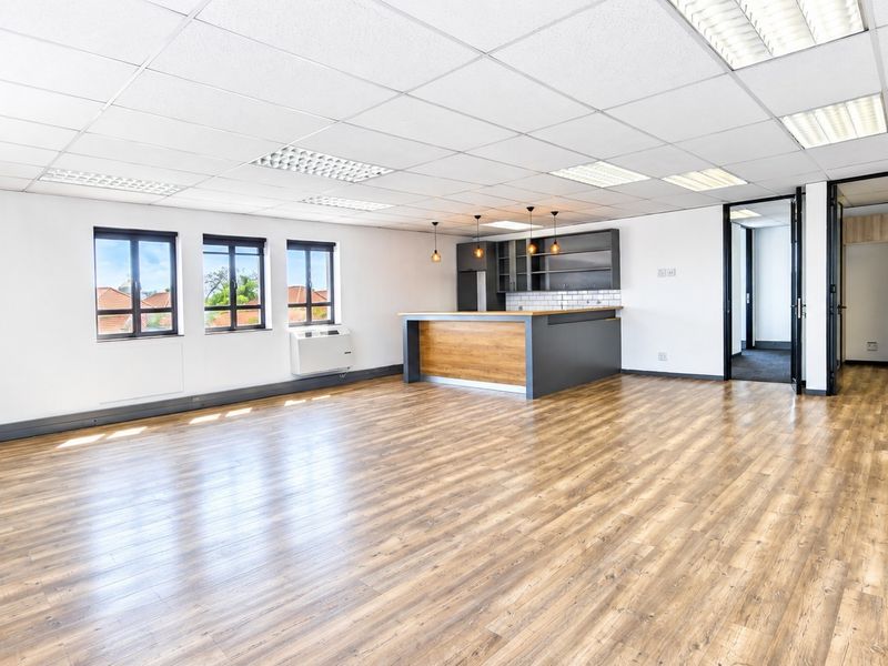 Spacious open-plan office area featuring modern laminate flooring, a built-in kitchenette, suspended ceilings with recessed lighting, and large windows providing natural light.