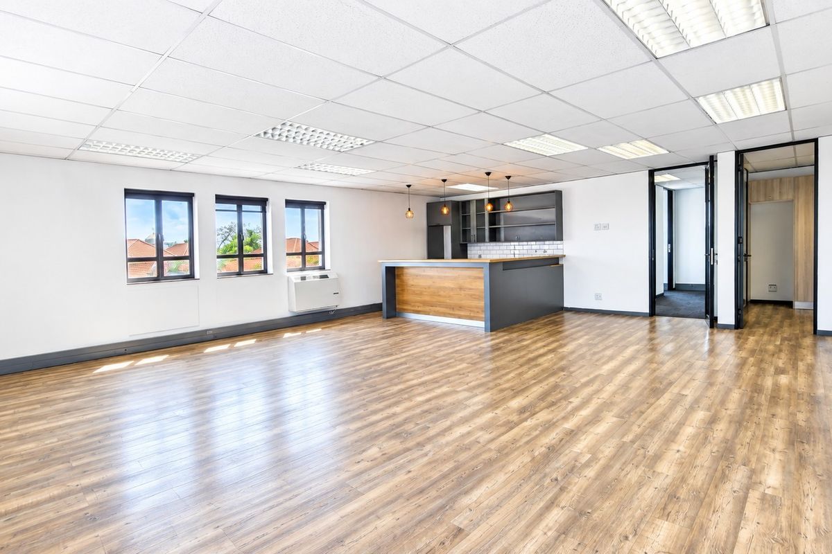 Spacious open-plan office area featuring modern laminate flooring, a built-in kitchenette, suspended ceilings with recessed lighting, and large windows providing natural light.