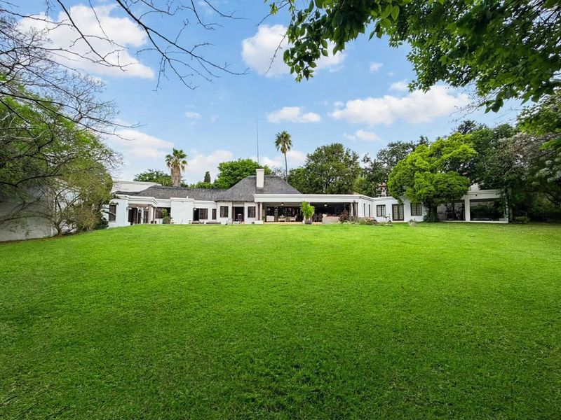 A spacious rear view of the property featuring a large manicured lawn, mature garden, and a covered patio area attached to the main residence.