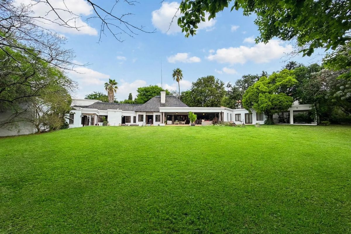 A spacious rear view of the property featuring a large manicured lawn, mature garden, and a covered patio area attached to the main residence.