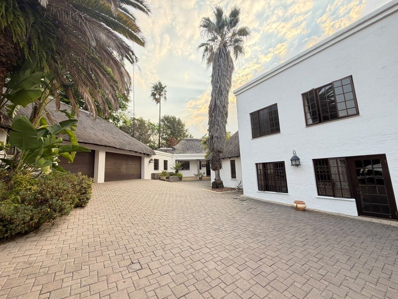 Spacious paved front courtyard featuring a multi-level residence with a combination of white textured walls, dark window frames, and traditional thatched roofing.