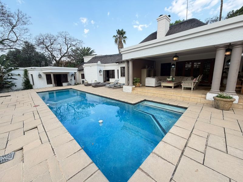 Spacious outdoor pool area featuring a large swimming pool, stone-paved deck, and a covered patio with a thatched roof and classic columns.