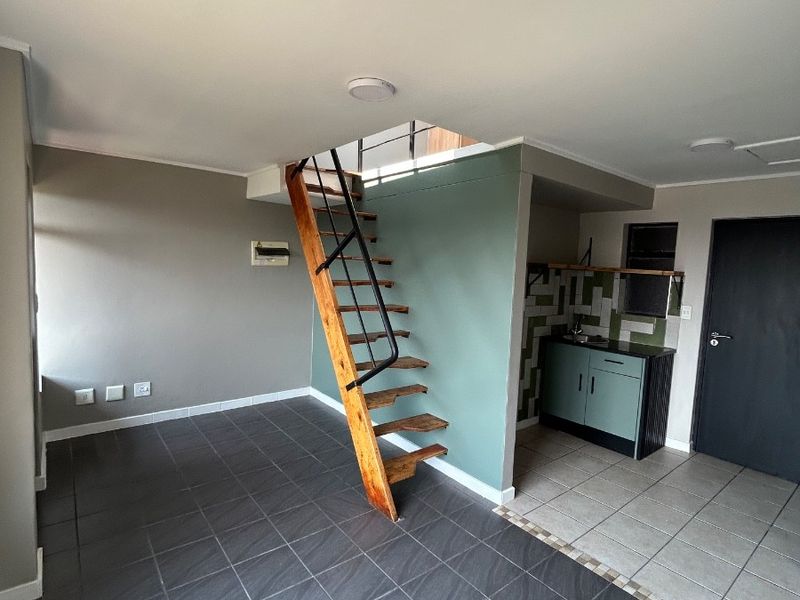 Open plan living area featuring dark floor tiles, a wooden staircase, and a compact kitchenette with built-in cabinetry and tiled backsplash.