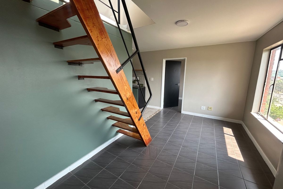 An open-plan living area featuring a distinctive open-tread wooden staircase, dark tiled flooring, and neutral wall finishes in a bright, vacant space.