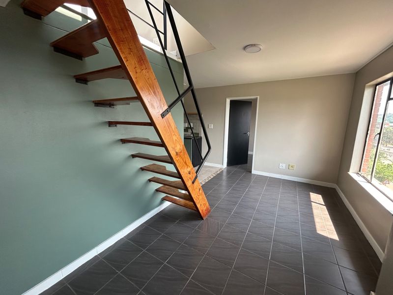 An open-plan living area featuring a distinctive open-tread wooden staircase, dark tiled flooring, and neutral wall finishes in a bright, vacant space.