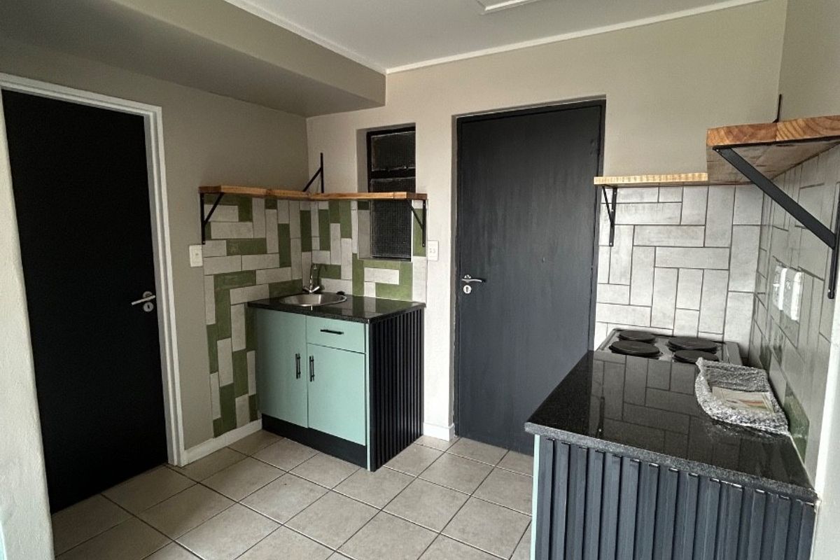 A compact, modern kitchen featuring built-in cabinetry, granite countertops, a tiled backsplash, and an electric hob, finished with neutral ceramic floor tiles.