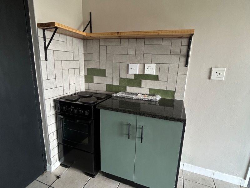 Compact kitchen area featuring a freestanding stove, sage green base cabinetry with granite countertop, and a decorative tiled backsplash with floating shelving.