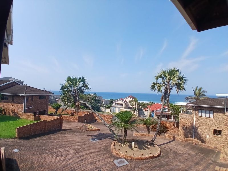 An elevated view overlooking a paved residential driveway, brick retaining walls, and landscaped garden beds with a scenic ocean backdrop.