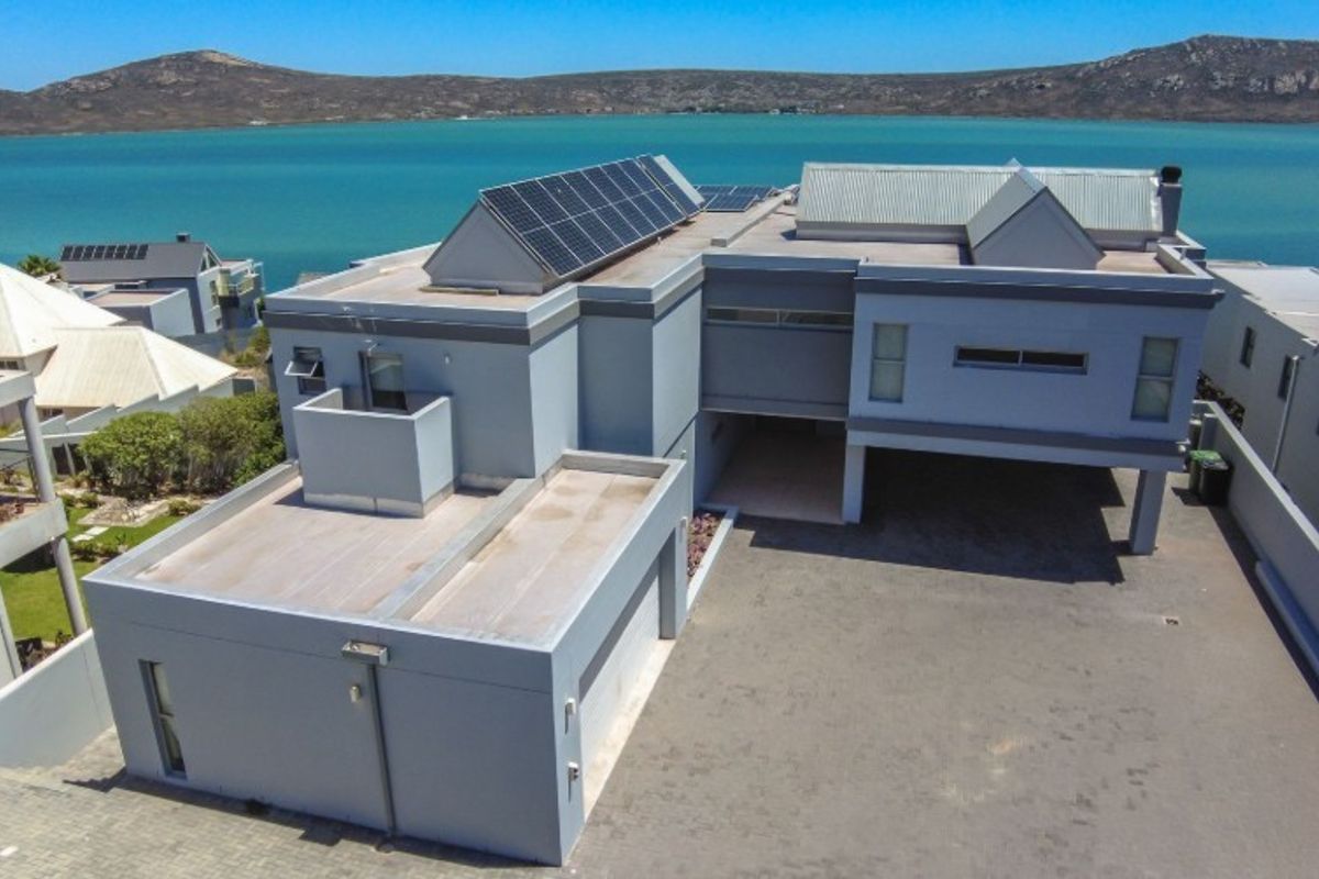 Aerial view of a modern multi-level residence featuring a paved driveway, integrated garage, and rooftop solar panels, set against a scenic coastal backdrop.