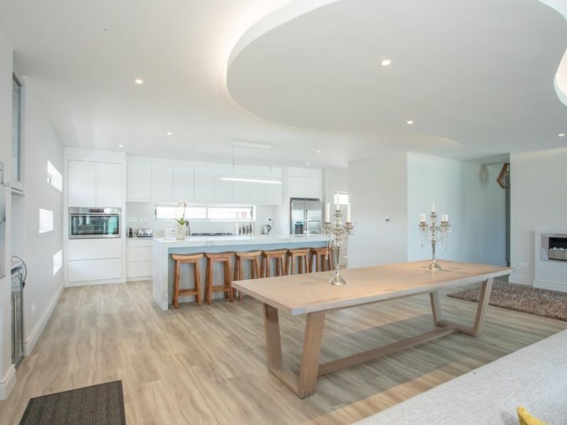 Modern open-plan living and kitchen area featuring a unique curved bulkhead ceiling, sleek white cabinetry, stone island bench, and a built-in fireplace.