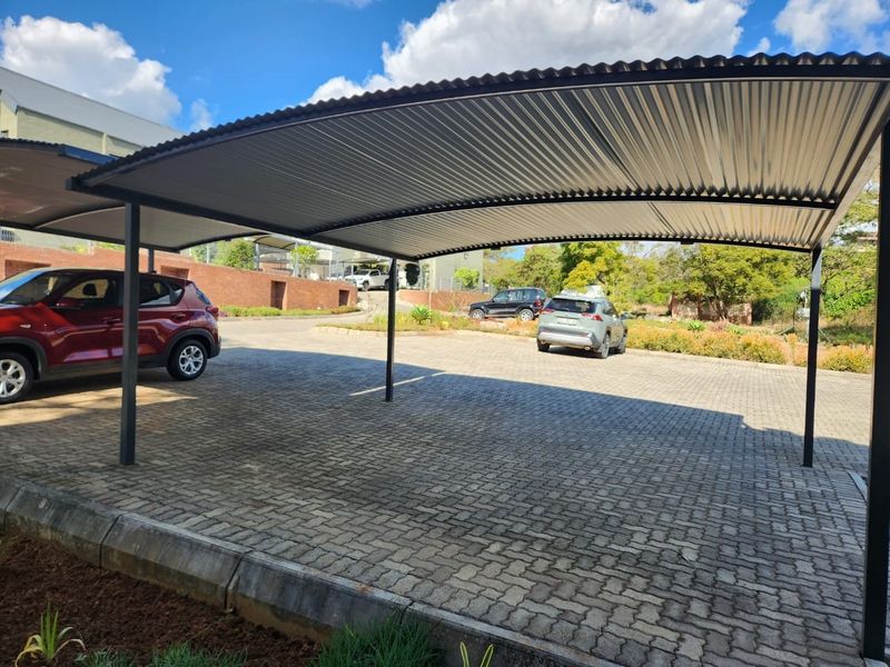 A covered carport structure featuring a corrugated metal roof supported by steel posts, situated over a paved driveway area.