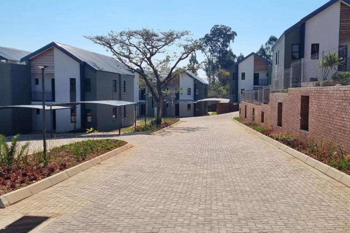 A view of the paved driveway and access road within a modern townhouse complex, featuring landscaped garden beds and contemporary building facades.