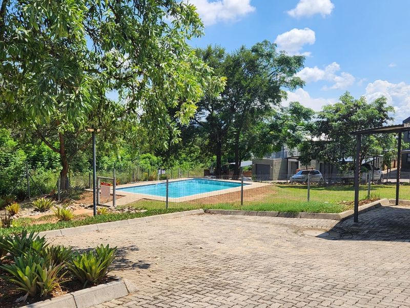 A communal outdoor area featuring a swimming pool, paved driveway, and carport structure within a landscaped residential setting.