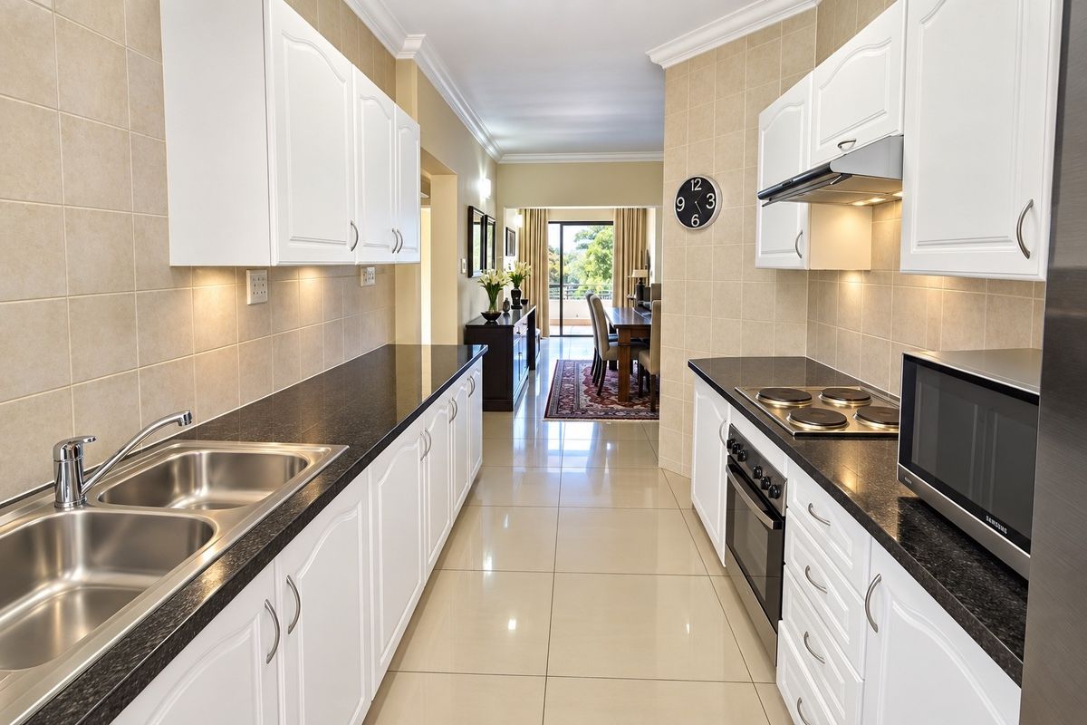 A well-maintained galley kitchen featuring white cabinetry, granite countertops, built-in oven, hob, and extractor fan, leading through to an open-plan dining area.