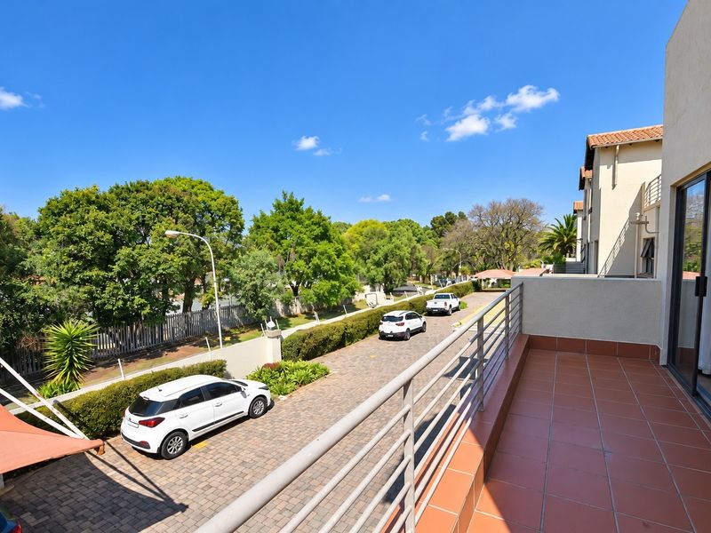 A spacious elevated balcony featuring tiled flooring and a metal balustrade, offering an open view of the complex driveway and surrounding greenery.
