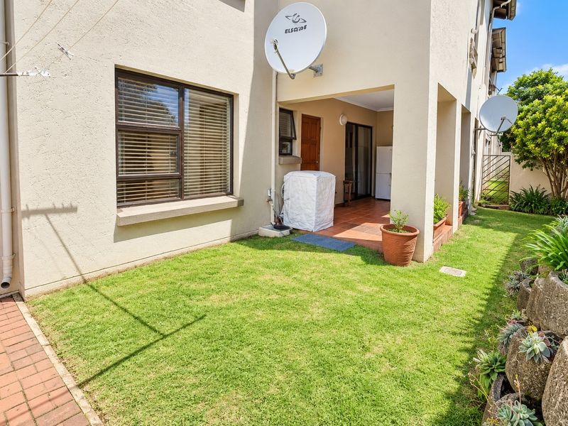 Rear exterior view of the townhouse featuring a covered patio, small private lawn, brick paved walkway, and a tiered garden retaining wall.