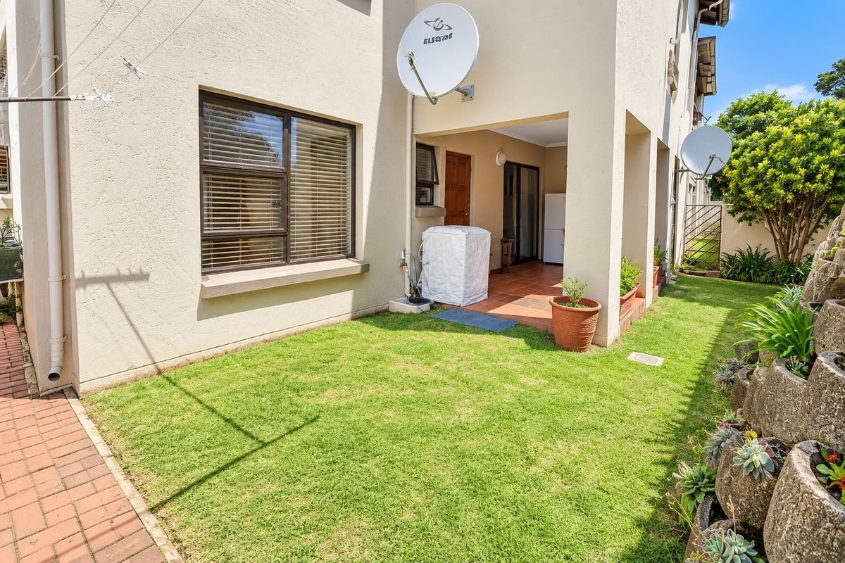 Rear exterior view of the townhouse featuring a covered patio, small private lawn, brick paved walkway, and a tiered garden retaining wall.