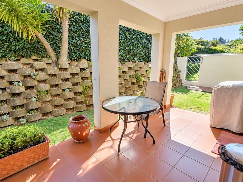 A covered patio featuring tiled flooring and concrete pillars, overlooking a landscaped garden with a tiered retaining wall.