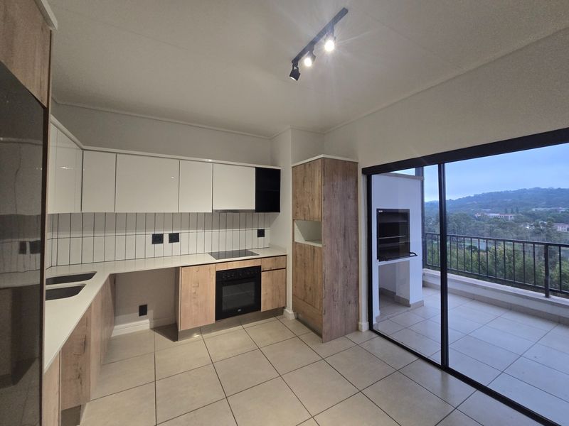 Modern open-plan kitchen featuring sleek cabinetry, integrated appliances, and tiled flooring, seamlessly connecting to a balcony with a built-in braai and scenic views.