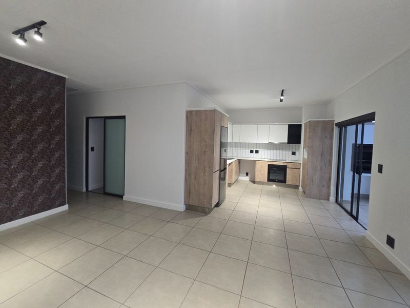 Spacious open-plan living area featuring a modern kitchen with built-in cabinetry, oven, and hob, leading to a bright, tiled living space.