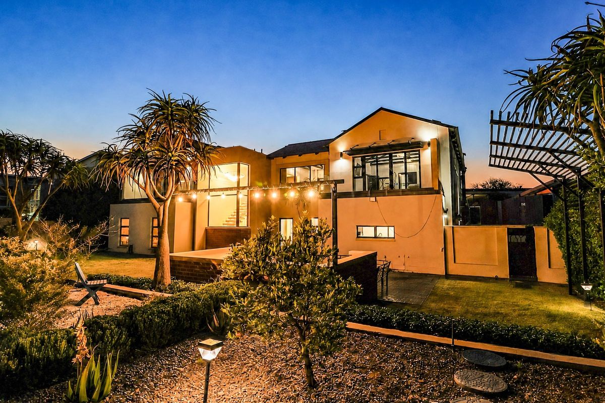 Rear view of the property featuring a landscaped garden, paved patio area, and exterior lighting, showcasing the home's architectural design at dusk.
