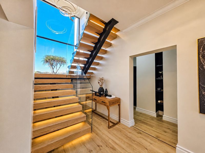 Modern entrance hall featuring a striking timber staircase with glass balustrade, integrated lighting, and a large window providing natural light.
