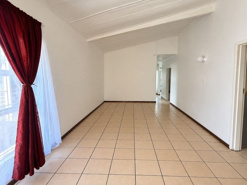 A spacious, empty living area featuring neutral tiled flooring, a vaulted ceiling, and natural light from a large window.