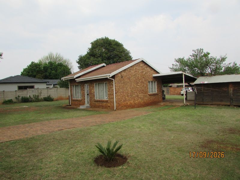 A detached face brick house featuring a tiled roof, attached carport, and a paved driveway set on a spacious grassed plot.
