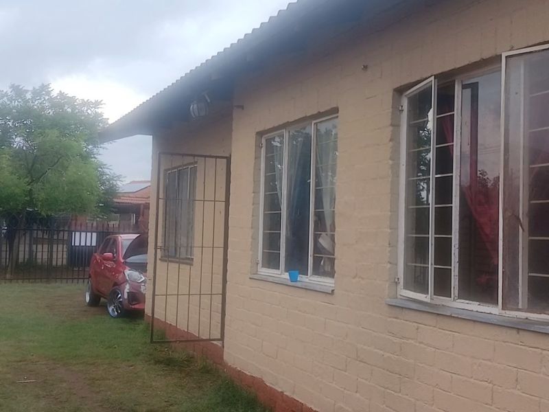 Rear view of a single-story brick house featuring security bars on windows and a corrugated metal roof, set within a garden area.