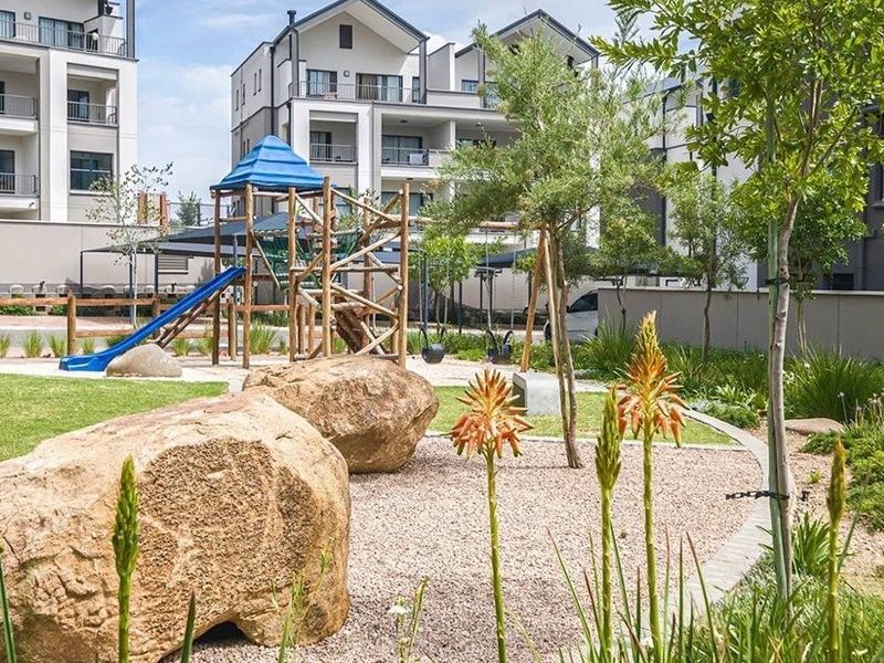 A well-maintained communal garden area featuring a children's playground, landscaped beds, and paved pathways within a modern apartment complex.