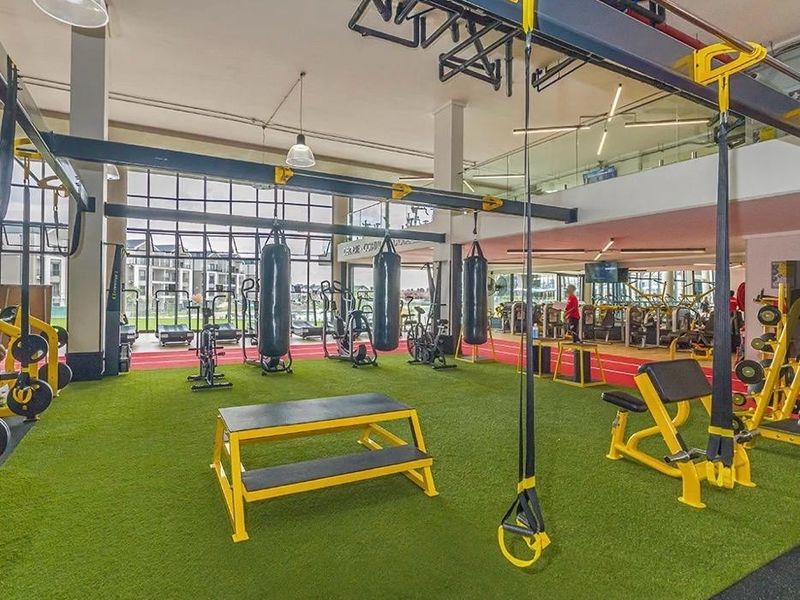 A modern, well-equipped apartment complex gym featuring artificial turf flooring, industrial-style exposed beams, and expansive windows providing ample natural light.