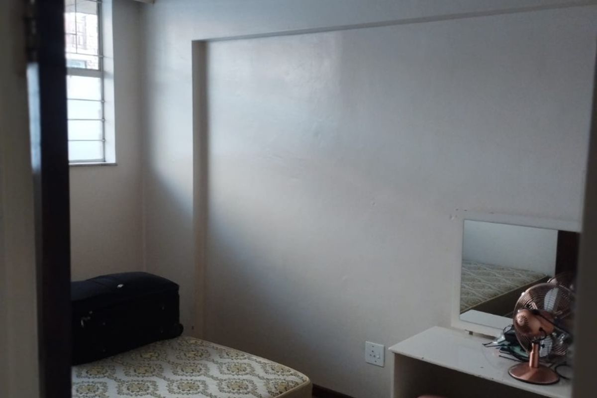 A compact bedroom featuring carpeted flooring, a window with security bars, and a simple ceiling light fixture.