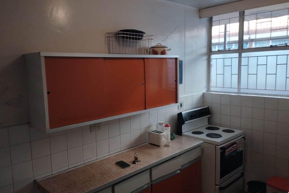 A compact kitchen featuring dated orange cabinetry, tiled backsplash, laminate countertops, and a freestanding electric stove with security bars on the window.