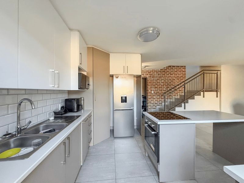 Modern kitchen featuring sleek cabinetry, a central island with built-in oven and cooktop, subway tile backsplash, and an adjacent staircase with exposed brick wall.