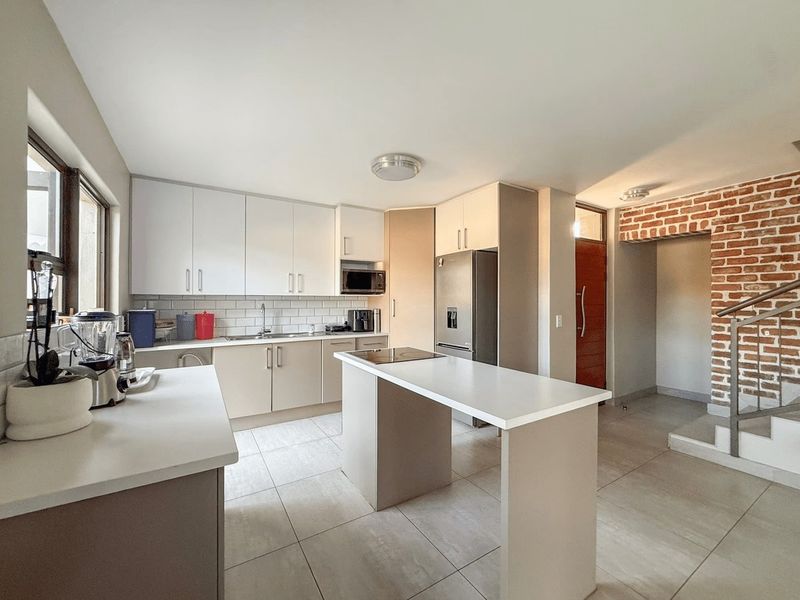 Modern kitchen featuring sleek cabinetry, a central island with cooktop, subway tile backsplash, and an attractive exposed brick feature wall near the staircase.