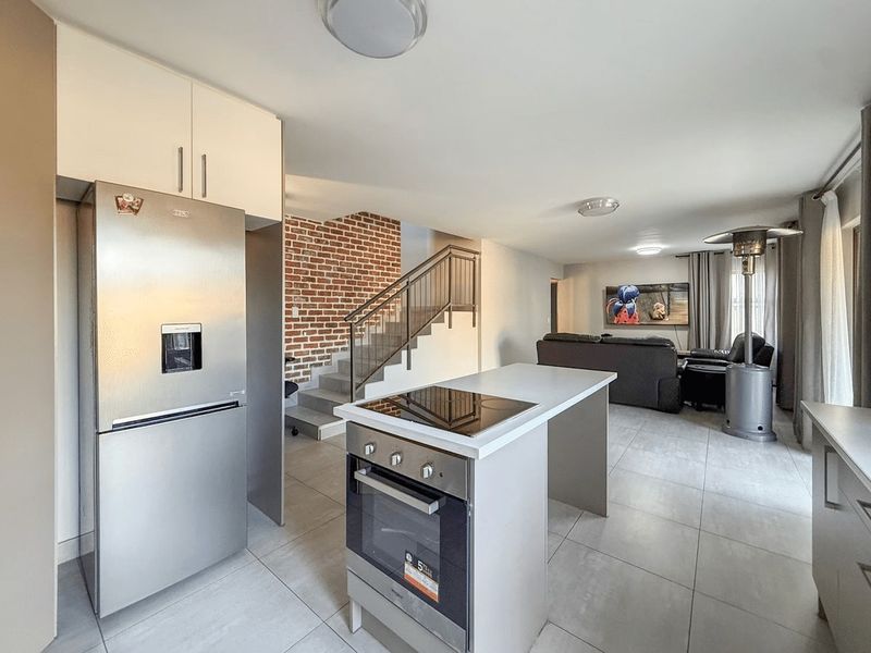 Modern open-plan living area featuring a kitchen island with built-in oven and cooktop, tiled floors, and a striking brick feature wall by the staircase.