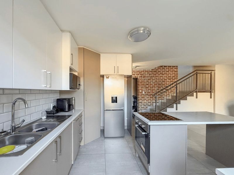 Modern kitchen featuring sleek cabinetry, subway tile backsplash, built-in oven and hob, adjacent to an exposed brick staircase.