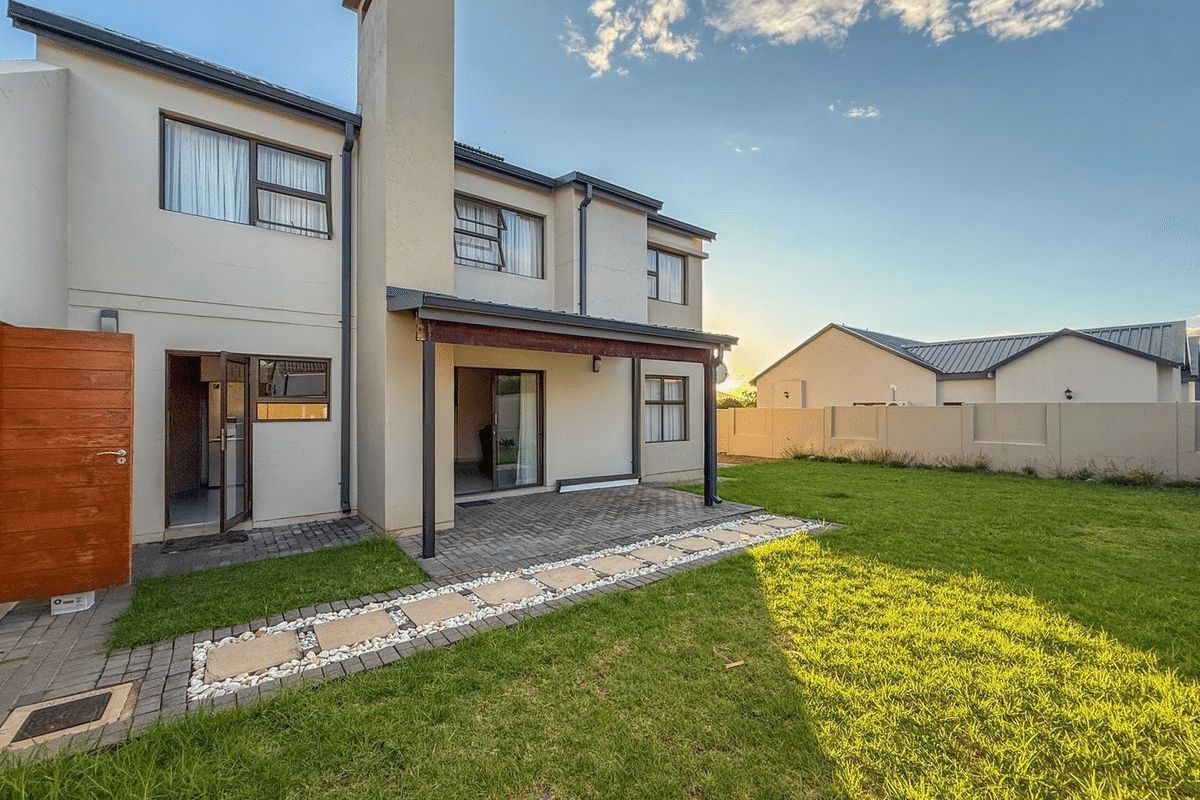 Rear view of the property featuring a covered patio, paved garden walkway, and a well-maintained lawn with boundary walling.