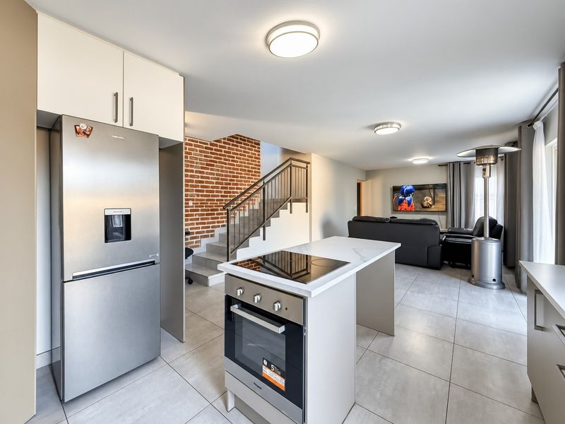 Modern open-plan living area featuring a kitchen island with built-in oven and cooktop, complemented by an exposed brick staircase and neutral tiled flooring.