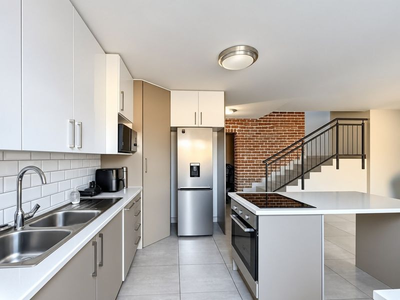 Modern kitchen featuring sleek cabinetry, a central island with integrated cooktop, subway tile backsplash, and an adjacent staircase with an exposed brick feature wall.