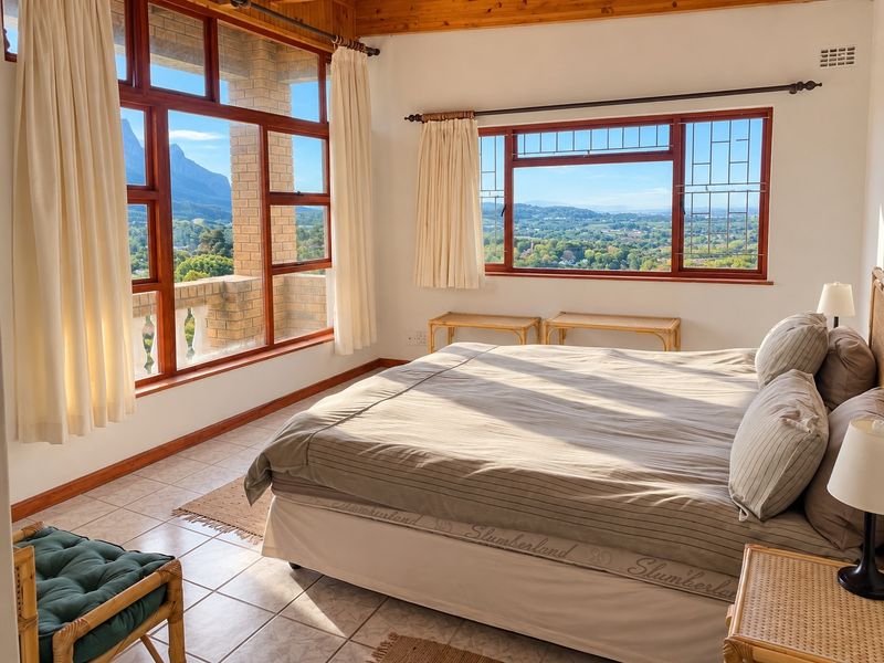 A bright bedroom featuring exposed timber ceiling beams, tiled flooring, and large timber-framed windows offering expansive mountain and landscape views.