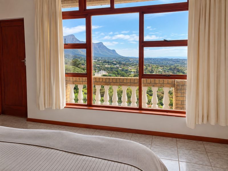 A bedroom featuring large timber-framed windows that offer expansive mountain and landscape views, complemented by a classic balustrade and tiled flooring.