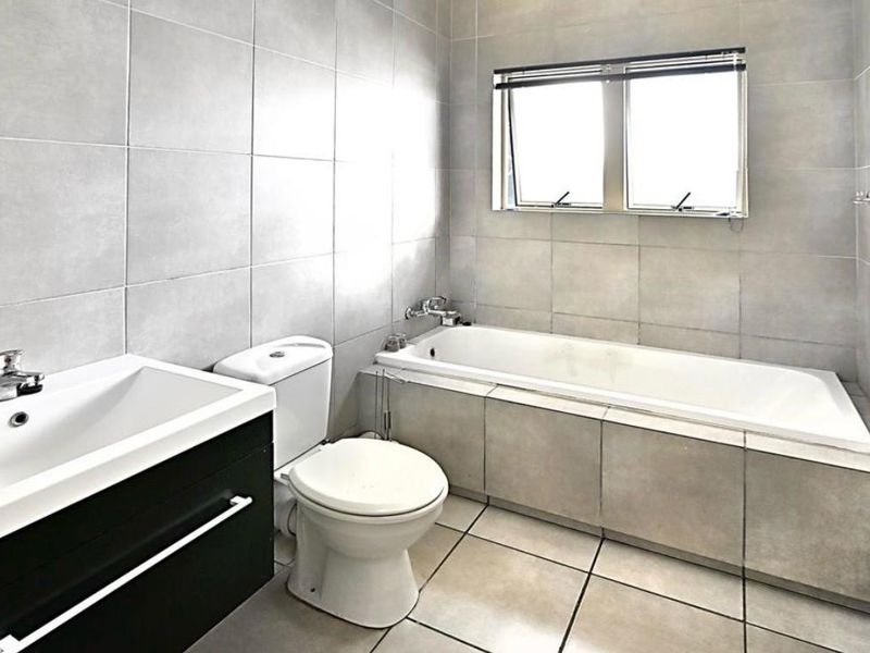 A modern, neutral-toned bathroom featuring a built-in vanity, a standard toilet, and a tiled bathtub area with large format wall and floor tiles.