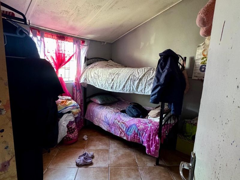 A compact bedroom featuring tiled flooring, a sloped ceiling, and security bars on the window, currently showing signs of wear and maintenance needs.